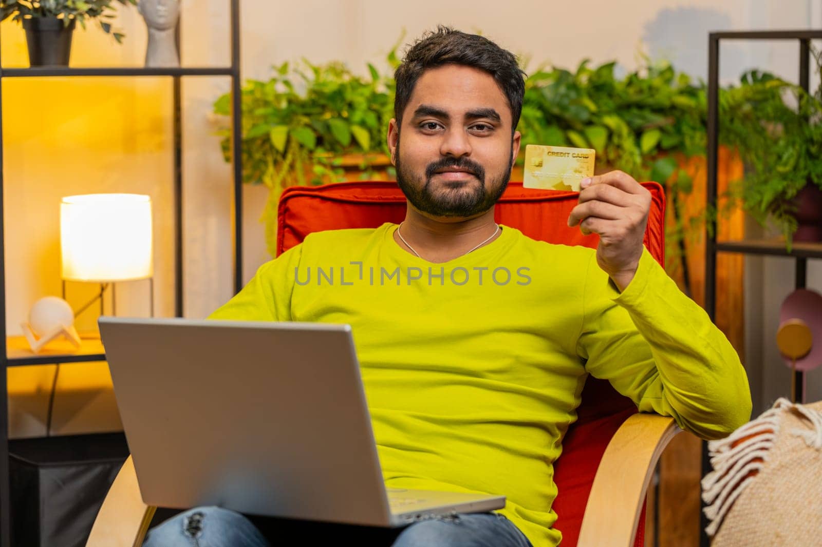 Indian man using credit bank plastic card and laptop computer while transferring money, purchases online shopping, order food delivery on internet at modern home room apartment. Guy in sits on chair