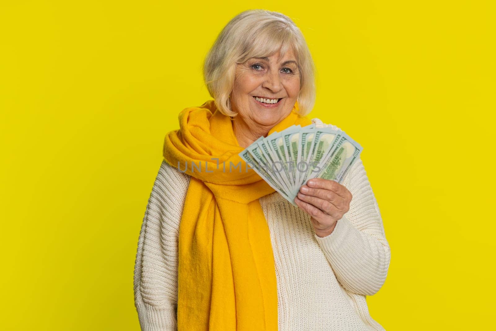 Rich happy senior old woman waving money dollar cash banknotes bills like a fan, success business career, lottery winner, big income wealth pension. Elderly grandmother pensioner on yellow background