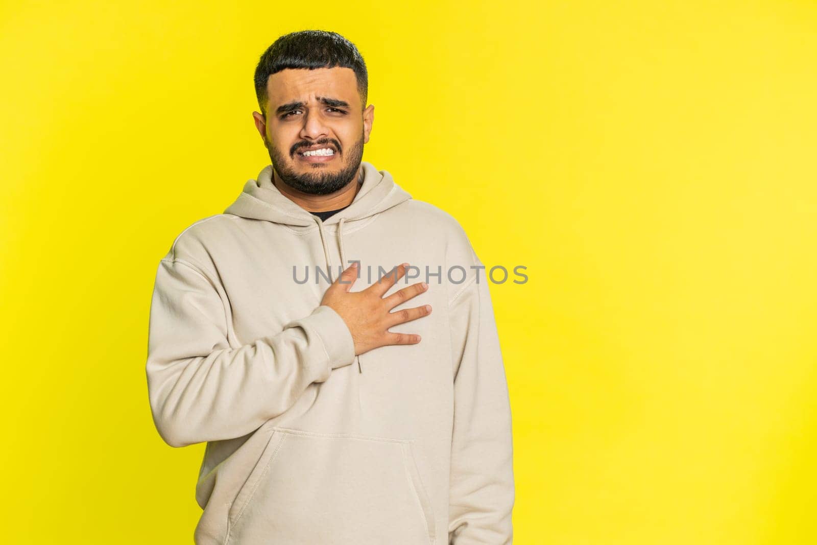 Stressed scared Indian man suffers from tension holding chest with heart pain attack risk of stroke pressure symptom, feeling bad sick ill. Arabian Hindu guy isolated on yellow background indoors