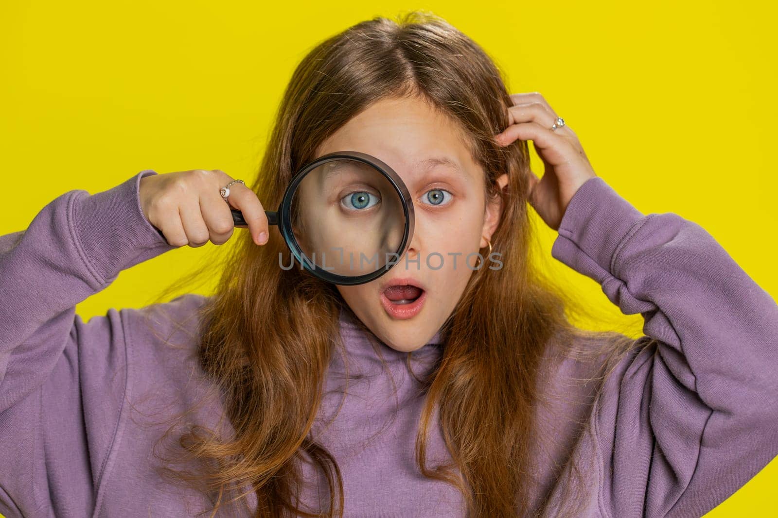 Investigator researcher scientist preteen child girl kid holding magnifying glass near face, looking into camera with big zoomed funny eye, searching, analyzing. School children on yellow background