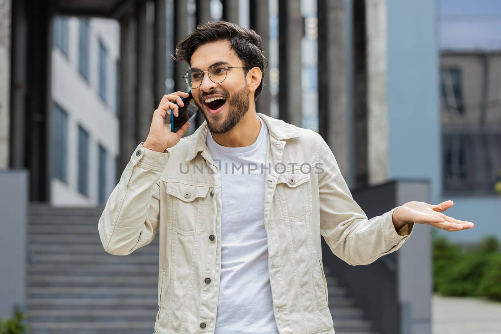 Phone call, good news, gossip. Happy surprised Indian man in pleasant conversation on smartphone, enjoying talking with friend outdoors. Arabian Hindu guy standing in city street. Town lifestyles