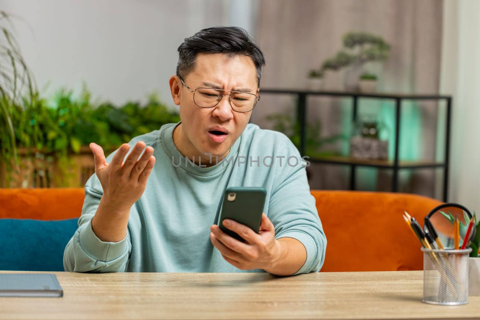 Sad displeased Chinese man use smartphone typing browsing, loses becoming surprised sudden lottery game results, bad news, fortune loss fail, deadline, virus. Mature Asian guy at home room on couch