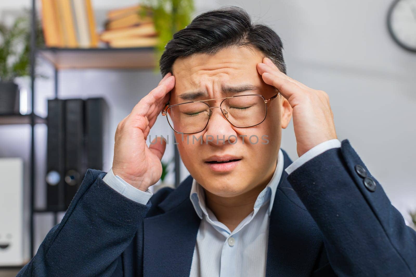 Close-up face of tired ill Asian businessman suffering from headache problem tension and migraine, stress at office desk. Mature freelancer broker worker man works on laptop notebook. Overworking