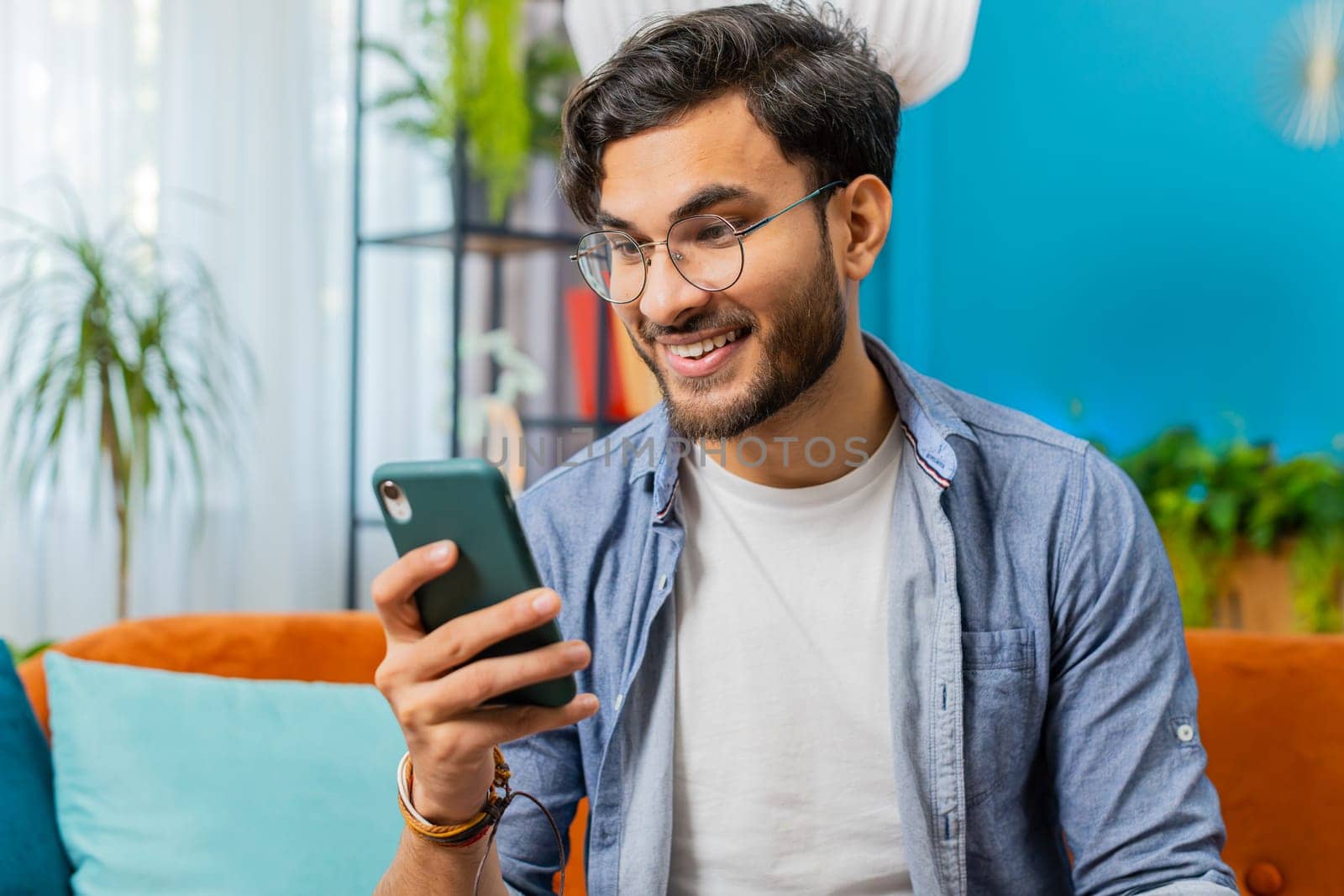 Indian man sits on couch uses mobile phone smiles at home living room apartment. Happy Arabian guy texting share messages content on smartphone social media applications online watching relax movie