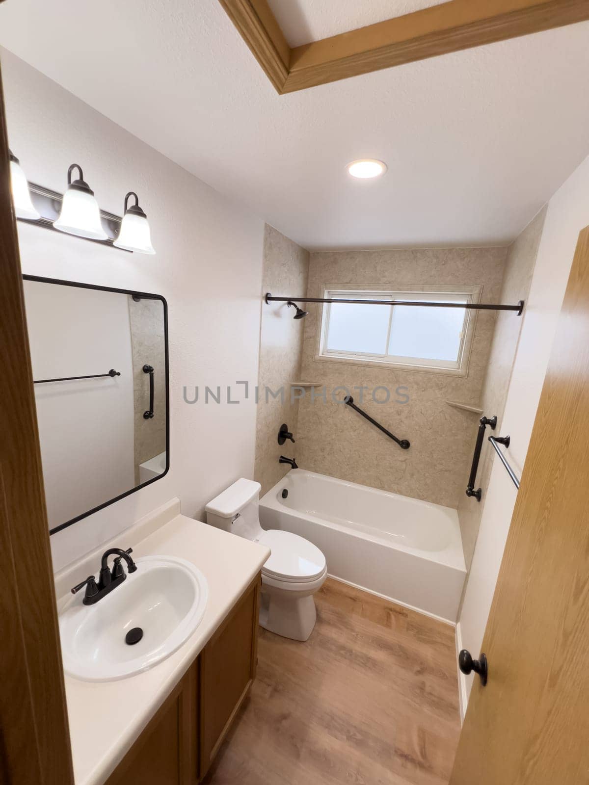 Broomfield, Colorado, USA-January 25, 2025-A modern bathroom featuring a white bathtub with beige tiled walls, a vanity with a white sink, and black fixtures. The bathroom includes a large mirror, a three-light vanity fixture, and a window allowing natural light. The wooden cabinets and light wood flooring complement the neutral color scheme. Black towel bars, a shower rod, and grab bars add a stylish contrast.
