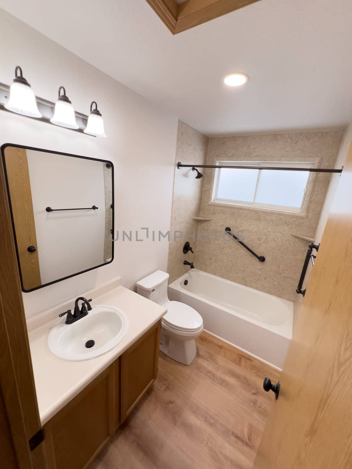 Broomfield, Colorado, USA-January 25, 2025-A modern bathroom featuring a white bathtub with beige tiled walls, a vanity with a white sink, and black fixtures. The bathroom includes a large mirror, a three-light vanity fixture, and a window allowing natural light. The wooden cabinets and light wood flooring complement the neutral color scheme. Black towel bars, a shower rod, and grab bars add a stylish contrast.