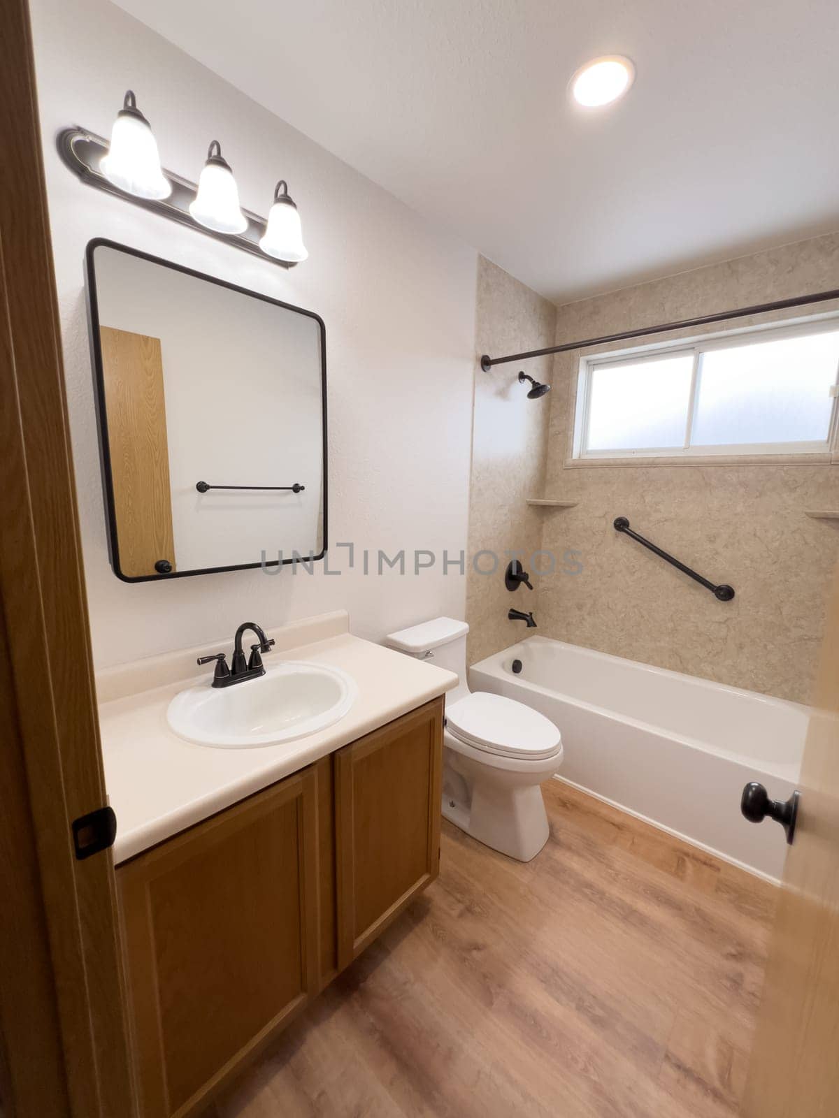 Broomfield, Colorado, USA-January 25, 2025-A modern bathroom featuring a white bathtub with beige tiled walls, a vanity with a white sink, and black fixtures. The bathroom includes a large mirror, a three-light vanity fixture, and a window allowing natural light. The wooden cabinets and light wood flooring complement the neutral color scheme. Black towel bars, a shower rod, and grab bars add a stylish contrast.