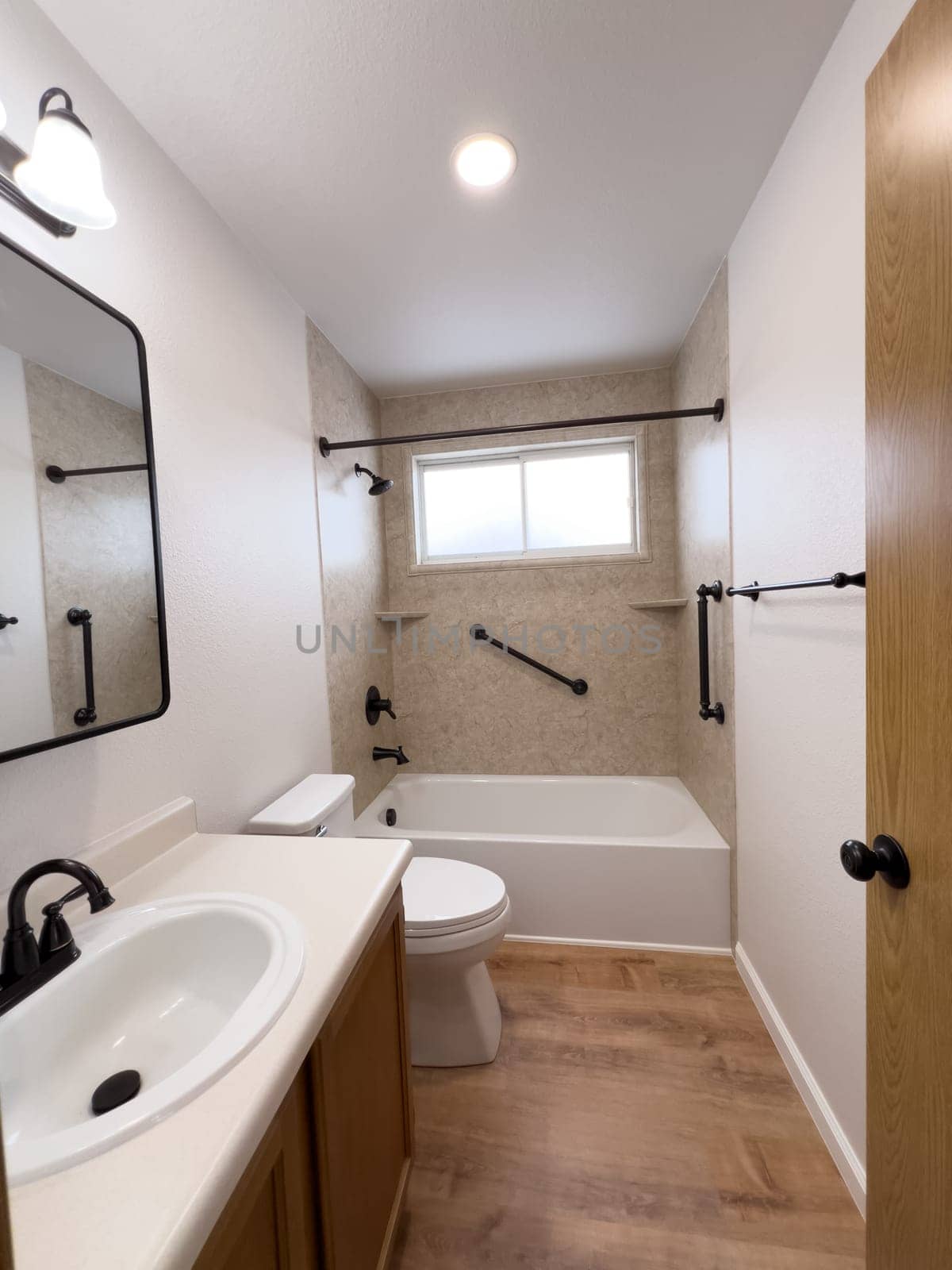 Broomfield, Colorado, USA-January 25, 2025-A modern bathroom featuring a white bathtub with beige tiled walls, a vanity with a white sink, and black fixtures. The bathroom includes a large mirror, a three-light vanity fixture, and a window allowing natural light. The wooden cabinets and light wood flooring complement the neutral color scheme. Black towel bars, a shower rod, and grab bars add a stylish contrast.
