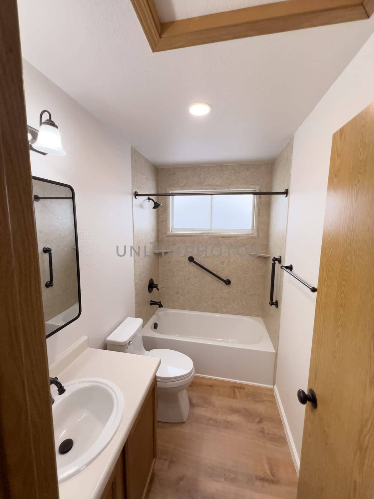 Broomfield, Colorado, USA-January 25, 2025-A modern bathroom featuring a white bathtub with beige tiled walls, a vanity with a white sink, and black fixtures. The bathroom includes a large mirror, a three-light vanity fixture, and a window allowing natural light. The wooden cabinets and light wood flooring complement the neutral color scheme. Black towel bars, a shower rod, and grab bars add a stylish contrast.