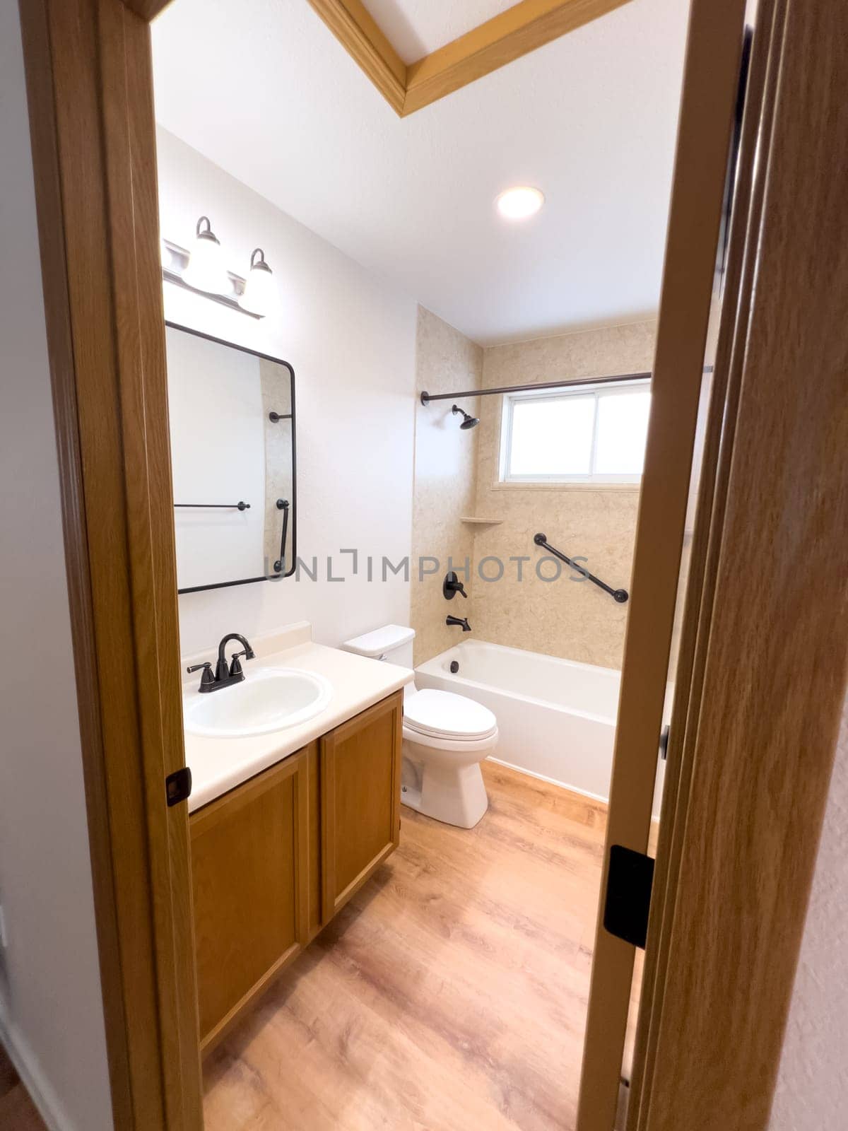 Broomfield, Colorado, USA-January 25, 2025-A modern bathroom featuring a white bathtub with beige tiled walls, a vanity with a white sink, and black fixtures. The bathroom includes a large mirror, a three-light vanity fixture, and a window allowing natural light. The wooden cabinets and light wood flooring complement the neutral color scheme. Black towel bars, a shower rod, and grab bars add a stylish contrast.