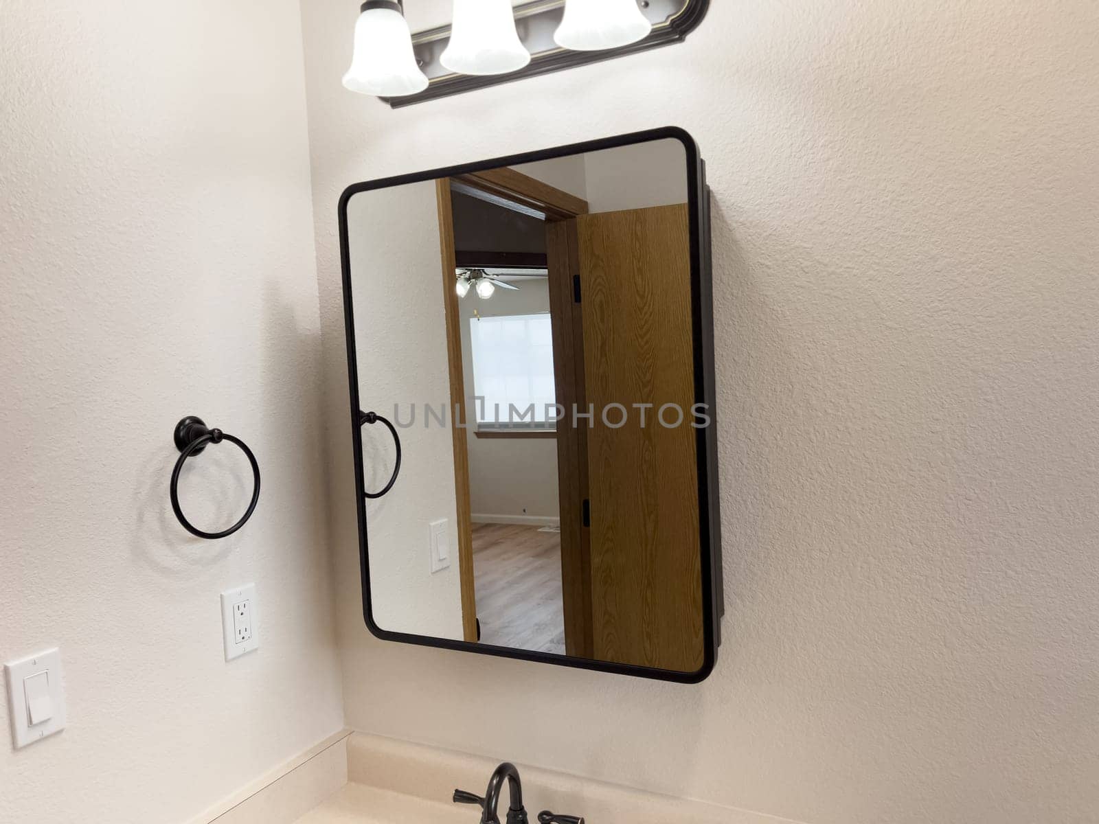 Broomfield, Colorado, USA-January 25, 2025-A stylish bathroom vanity featuring a large rectangular mirror with a black frame, a white sink with a black faucet, and a beige countertop. The vanity is complemented by a three-light fixture above the mirror, providing ample lighting. A black towel bar is mounted on the wall, enhancing the modern design. The bathroom has a clean, minimalist look with neutral tones and functional elements.
