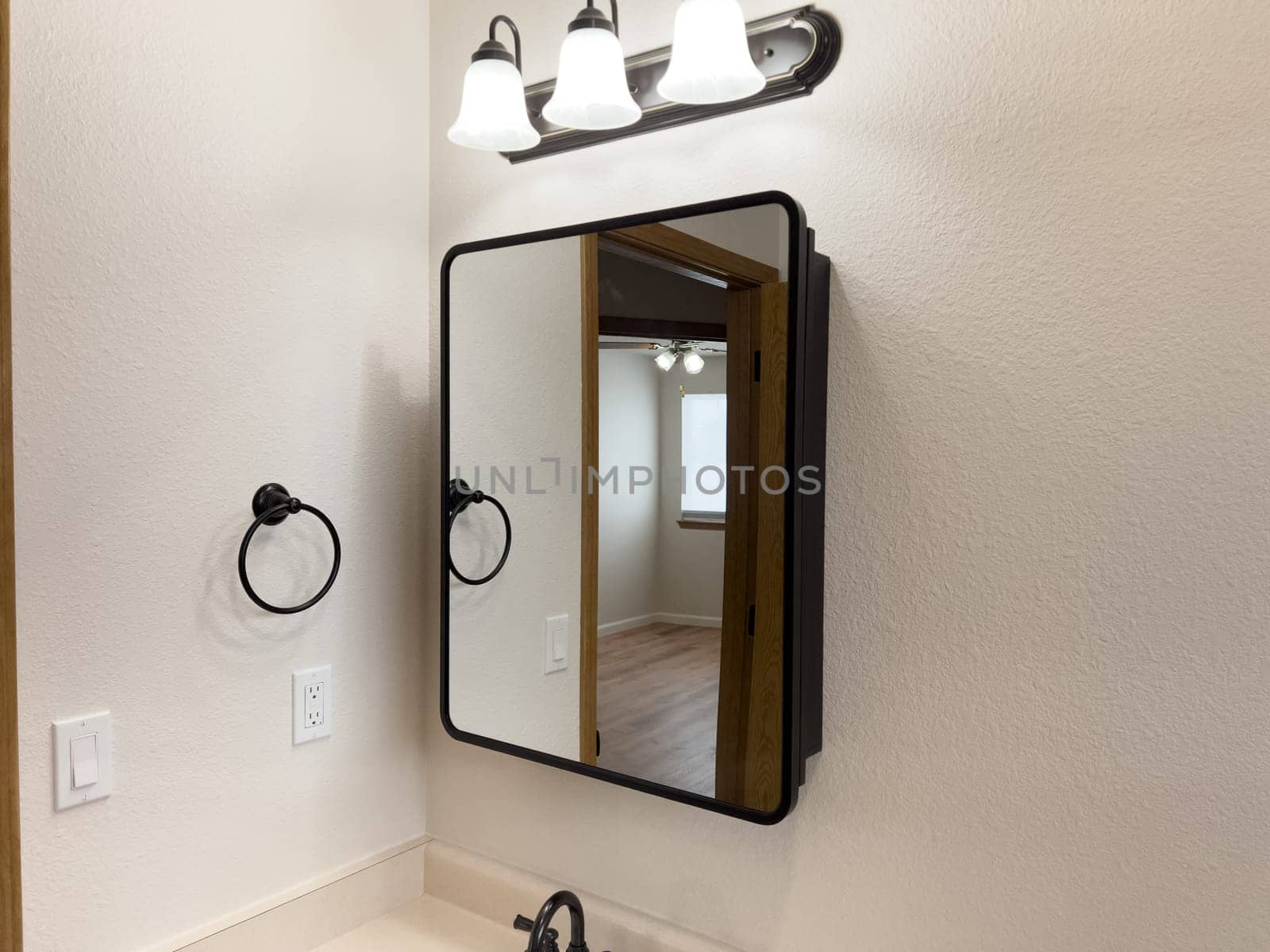 Broomfield, Colorado, USA-January 25, 2025-A stylish bathroom vanity featuring a large rectangular mirror with a black frame, a white sink with a black faucet, and a beige countertop. The vanity is complemented by a three-light fixture above the mirror, providing ample lighting. A black towel bar is mounted on the wall, enhancing the modern design. The bathroom has a clean, minimalist look with neutral tones and functional elements.