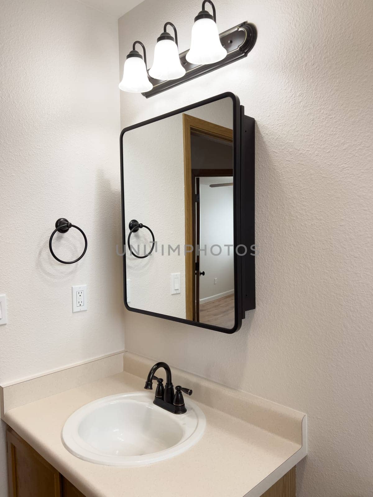 Broomfield, Colorado, USA-January 25, 2025-A stylish bathroom vanity featuring a large rectangular mirror with a black frame, a white sink with a black faucet, and a beige countertop. The vanity is complemented by a three-light fixture above the mirror, providing ample lighting. A black towel bar is mounted on the wall, enhancing the modern design. The bathroom has a clean, minimalist look with neutral tones and functional elements.