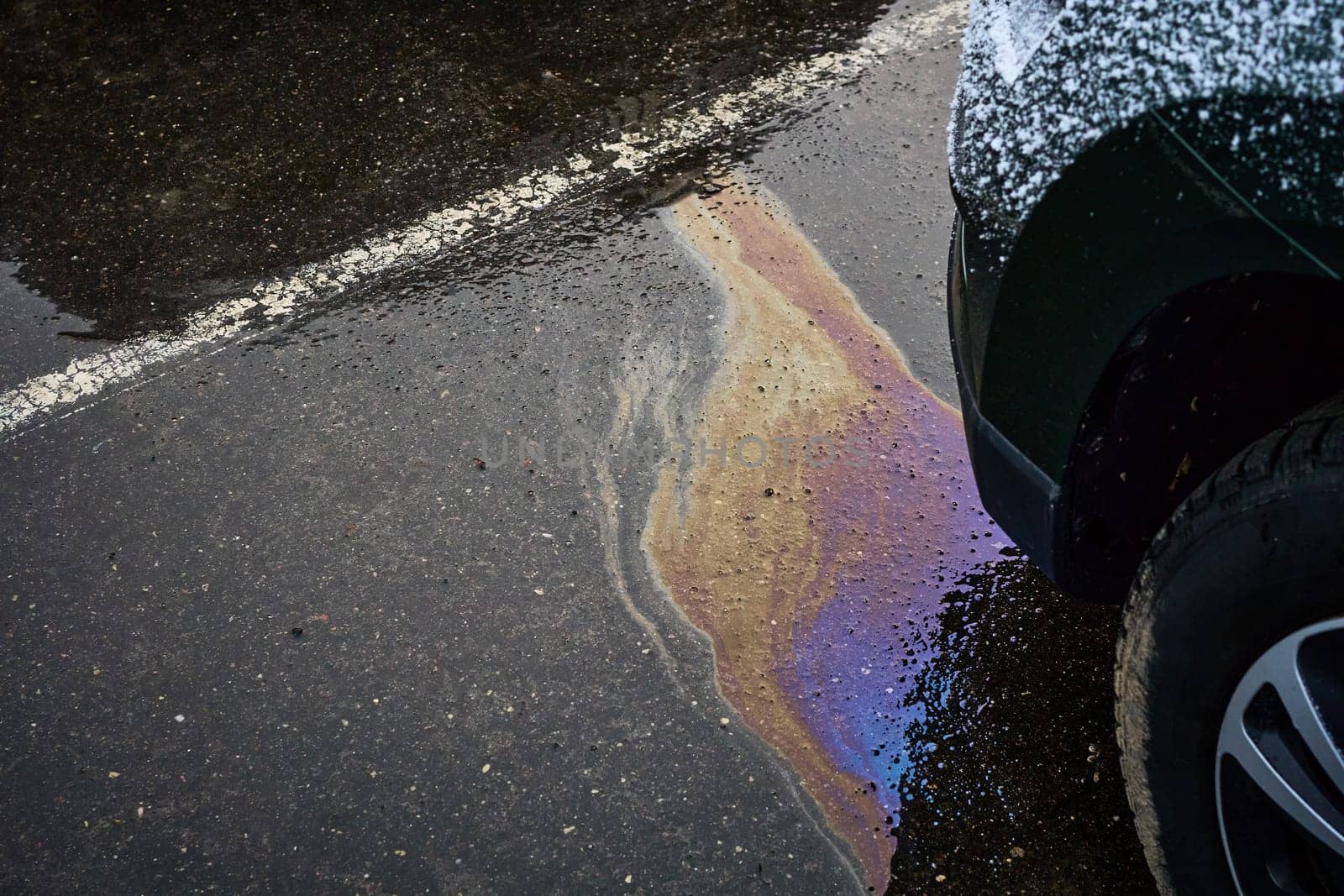 Colorful oil leak forming an iridescent sheen on the damp asphalt near a vehicle, drawing attention to pollution concerns.