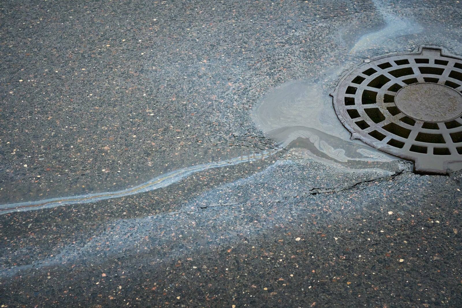 Gasoline or oil spill creating rainbow sheen near storm drain on asphalt road.