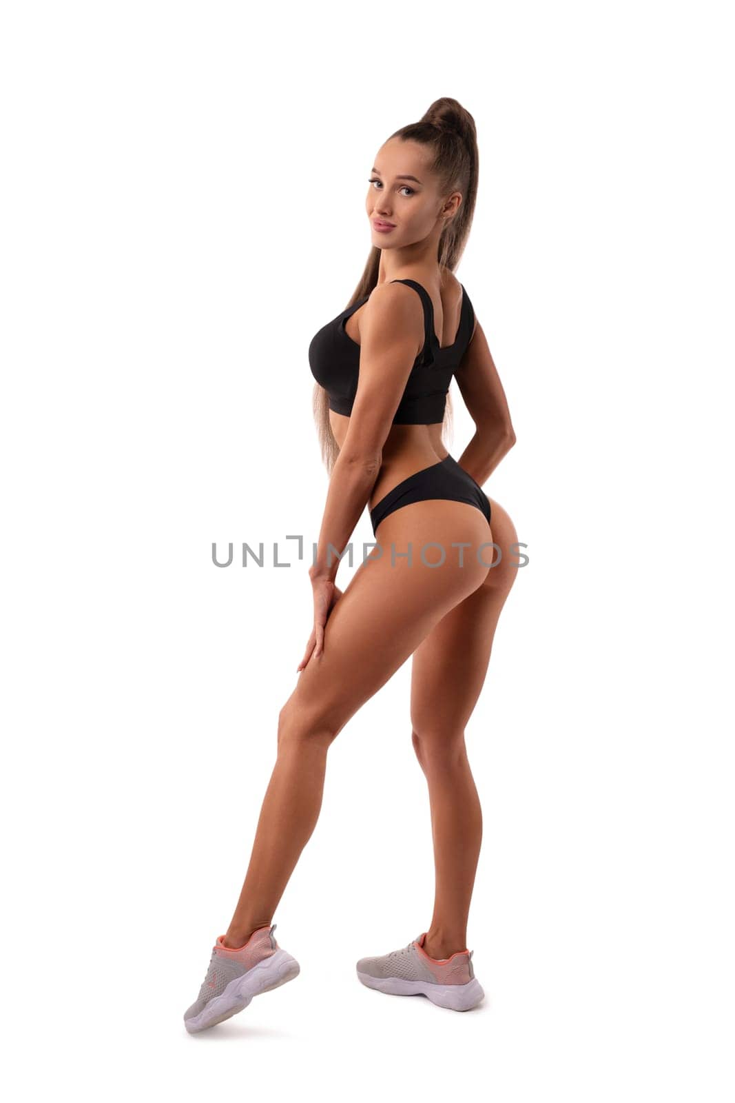 Side view of sexy slender female in sports bra and sneakers posing on white background in studio and looking at camera