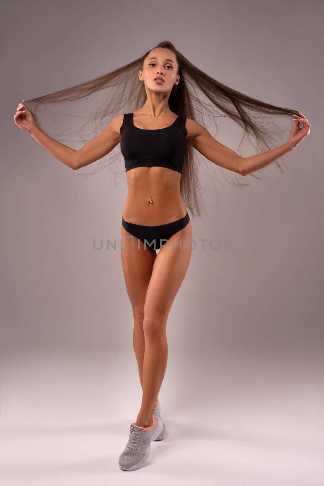 Seductive female in sports bra and panties standing in studio while touching long hair and looking at camera