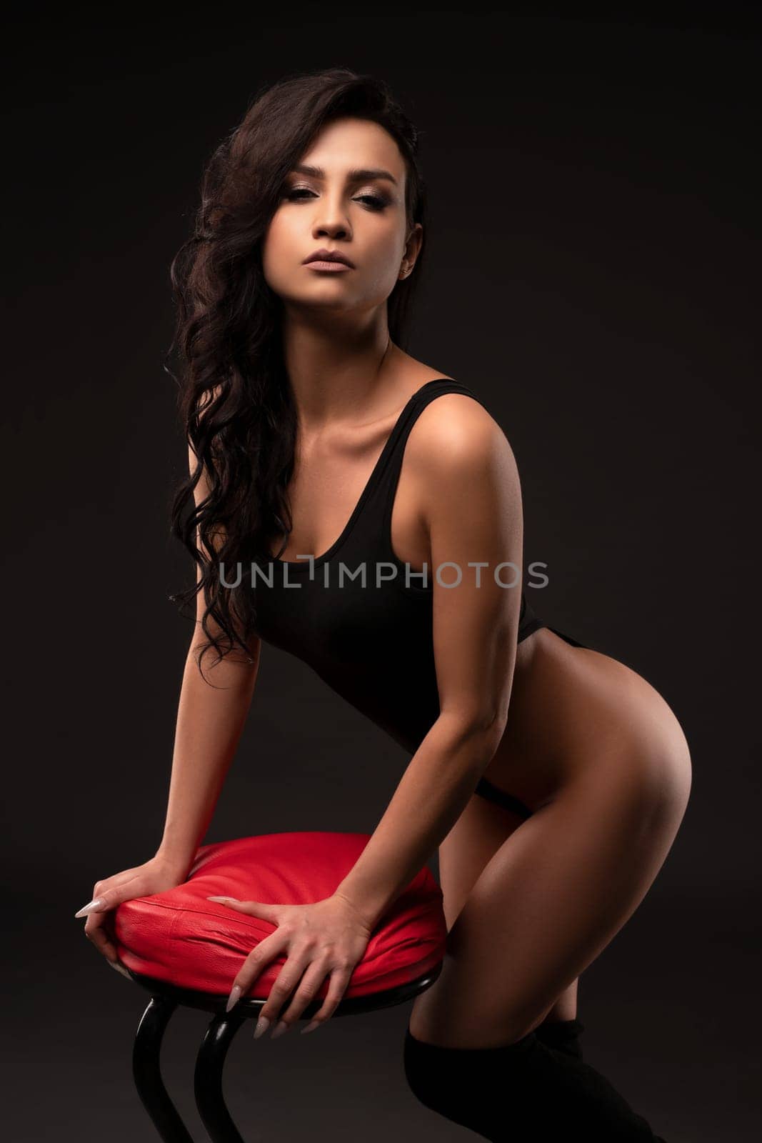 Side view of sexy female in black bodysuit leaning on stool while standing in studio on dark background and looking at camera
