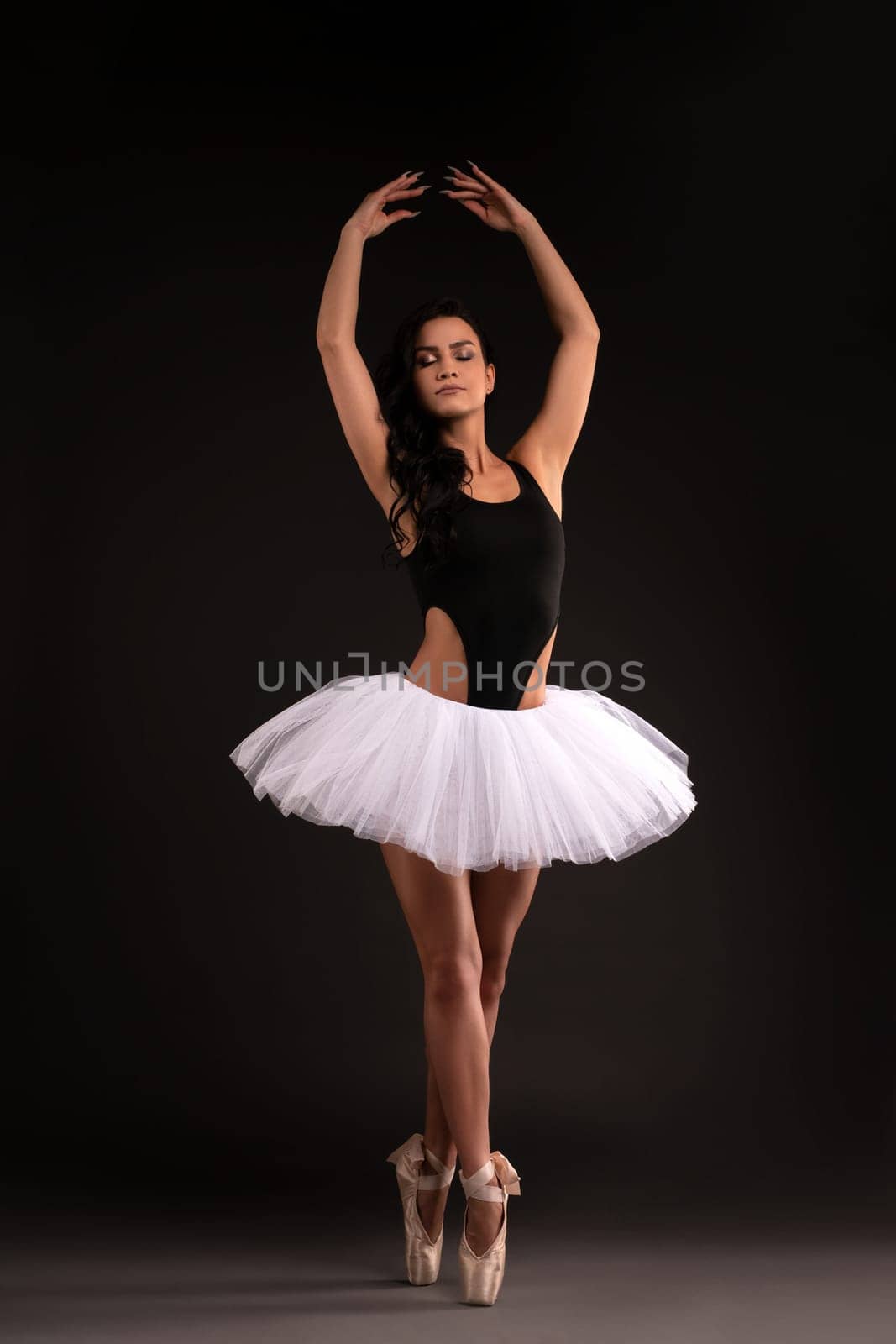 Silly female ballet dancer in tulle tutu skirt and pointe shoes standing in studio and shrugging shoulders while looking at camera