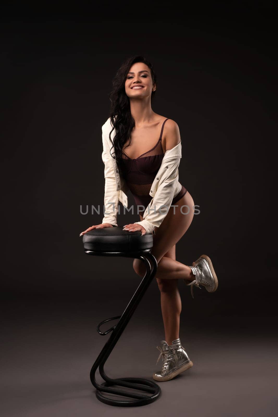 Slender cheerful female in underwear and sneakers leaning on stool while standing on dark background in studio and looking at camera