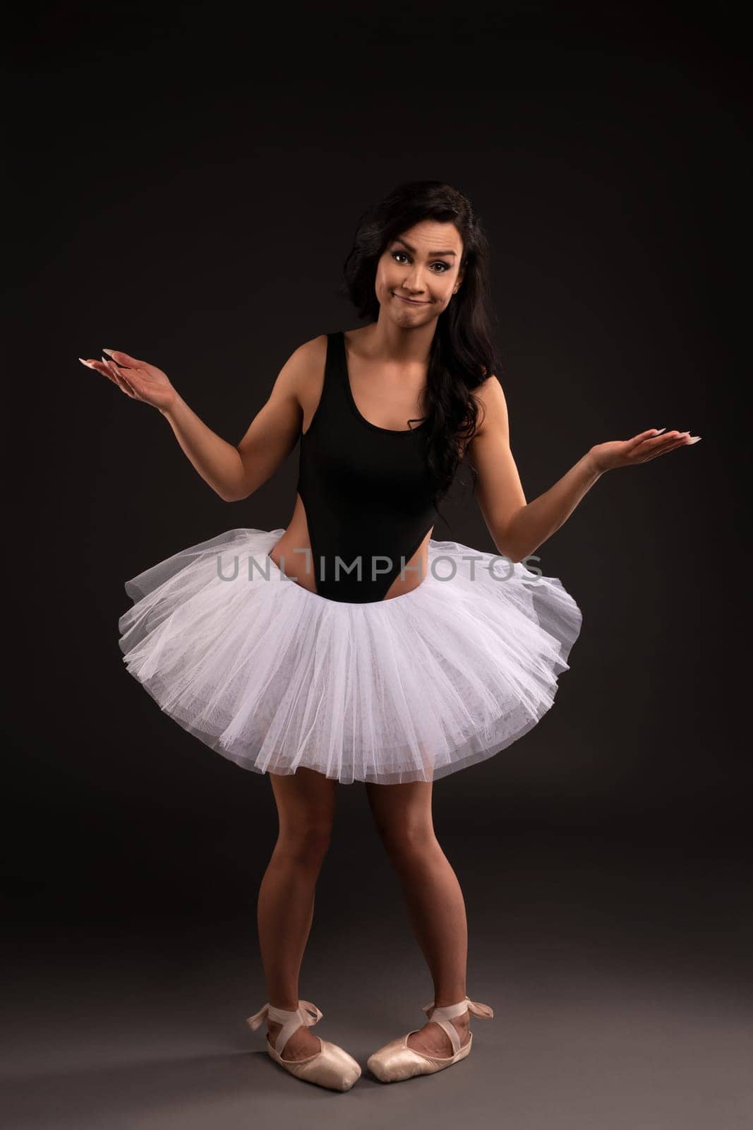 Silly female ballet dancer in tulle tutu skirt and pointe shoes standing in studio and shrugging shoulders while looking at camera