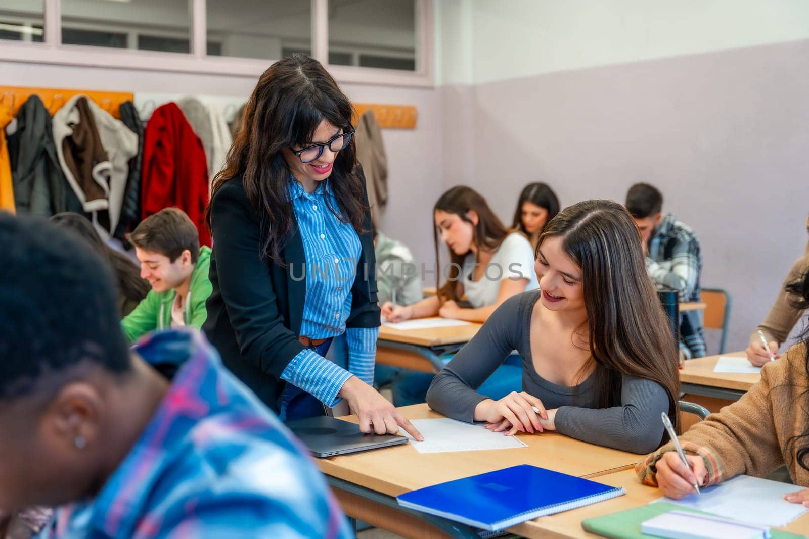 Female professor assisting a smiling student with her exam paper in a classroom full of diverse students taking a test