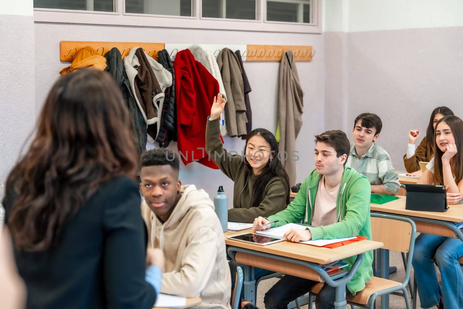 Multi ethnic group of high school students attending a lesson, a girl is raising her hand to ask a question to the teacher