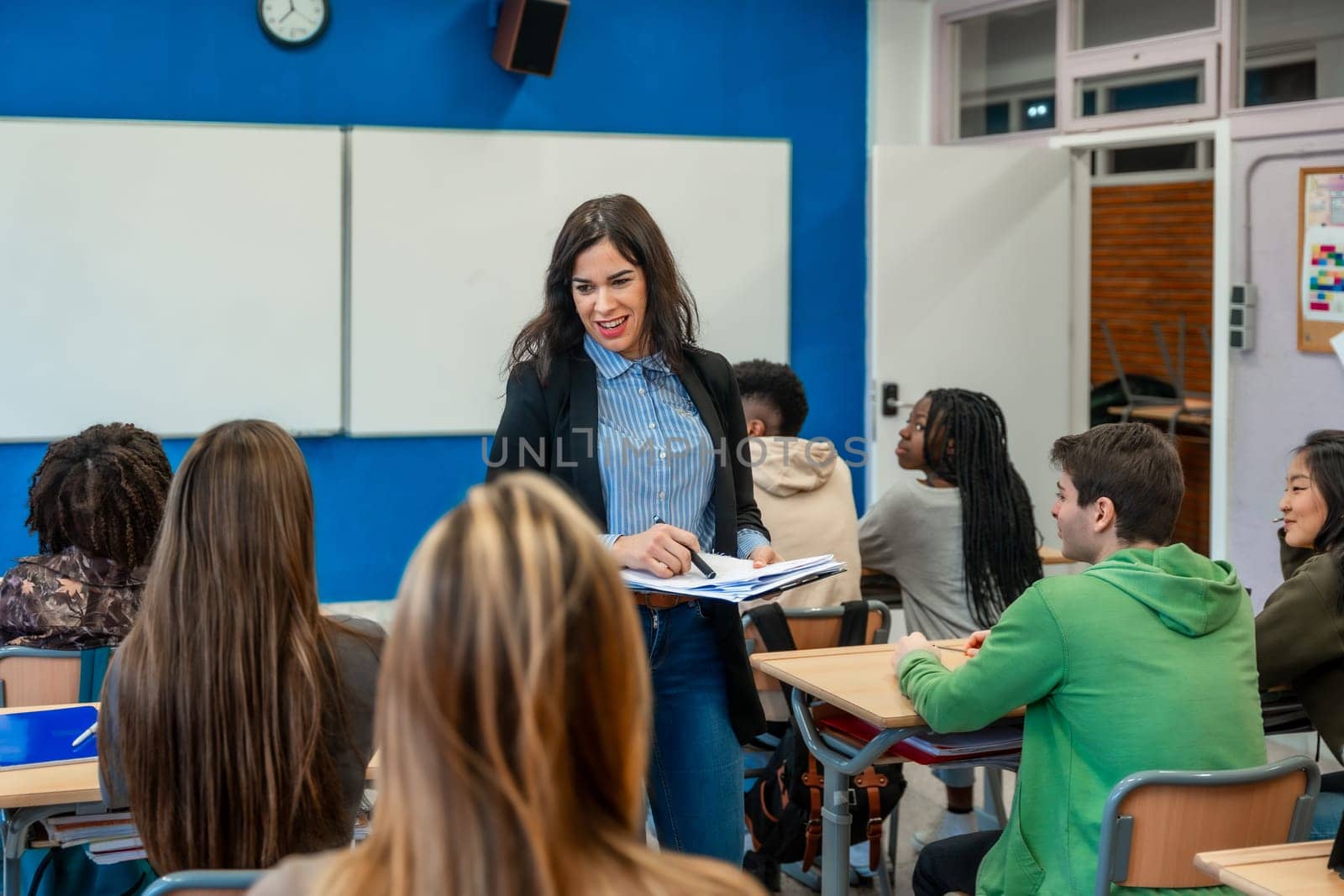 Smiling female teacher holding a notebook, interacting with her attentive high school students during a lesson in the classroom