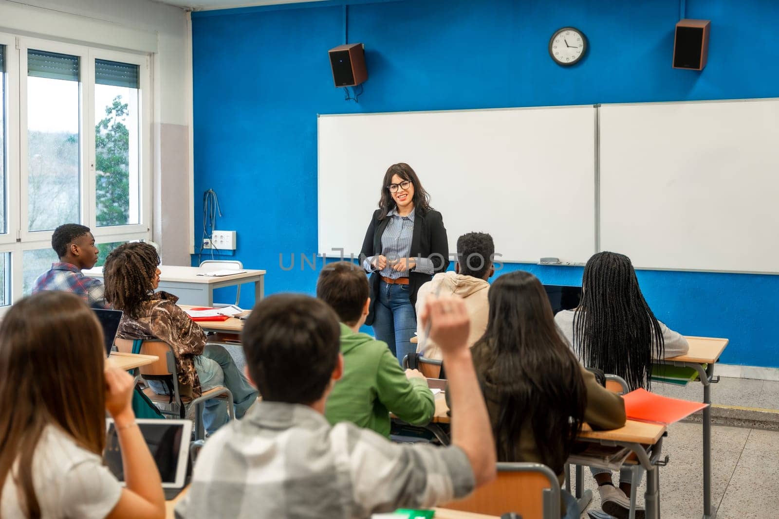 Multiethnic students raising hands during a lesson held by female teacher in high school classroom