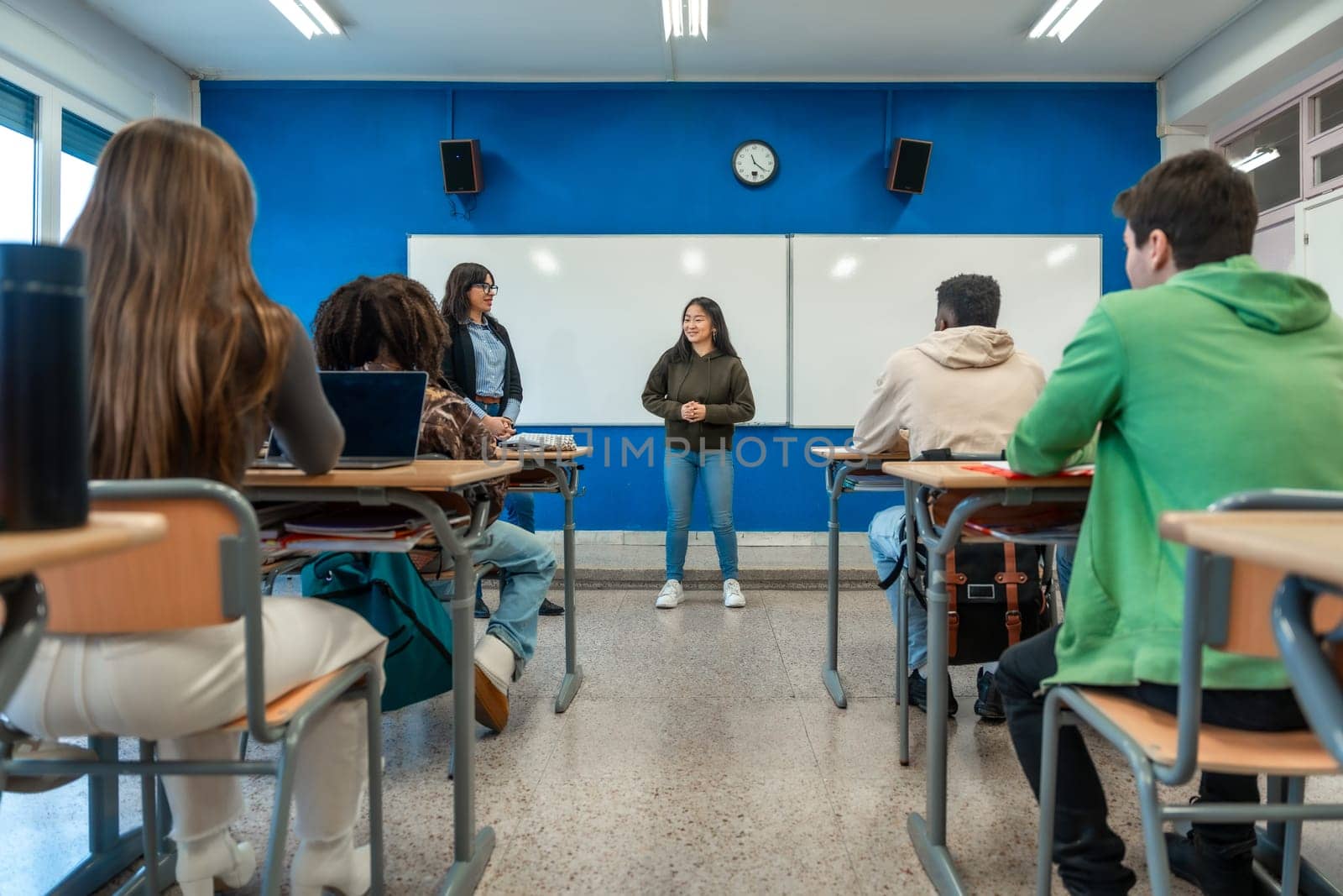 Teenager girl giving a presentation in a classroom while teacher and classmates listen to her
