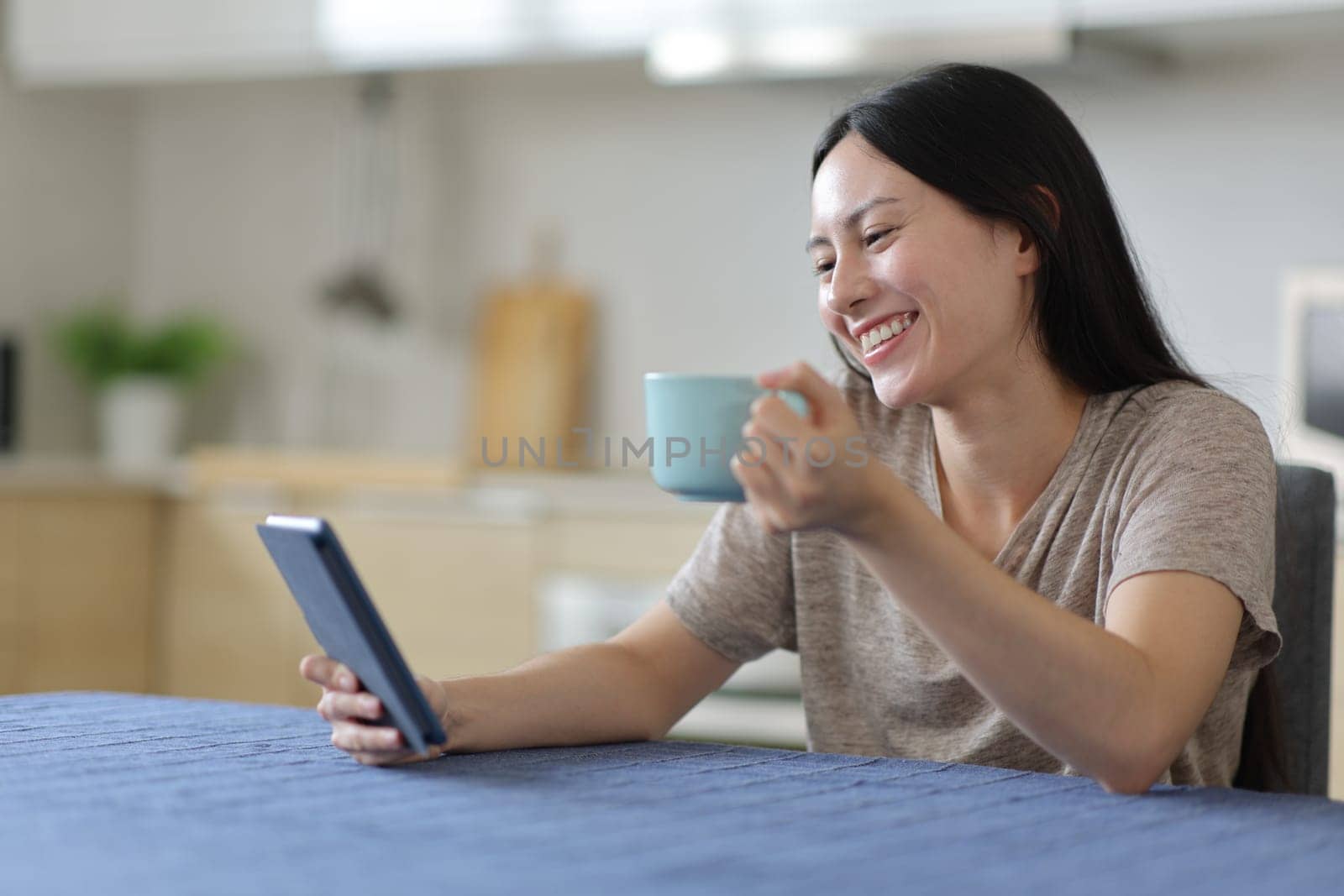 Happy asian woman reading ebook drinking coffee in the kitchen at home