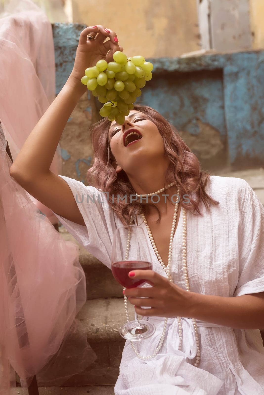 young woman eating grapes and drinking red wine. she is wearing a summer white dress. sitting on the steps of an old building. holding a bunch of grapes and a glass of wine, close-up