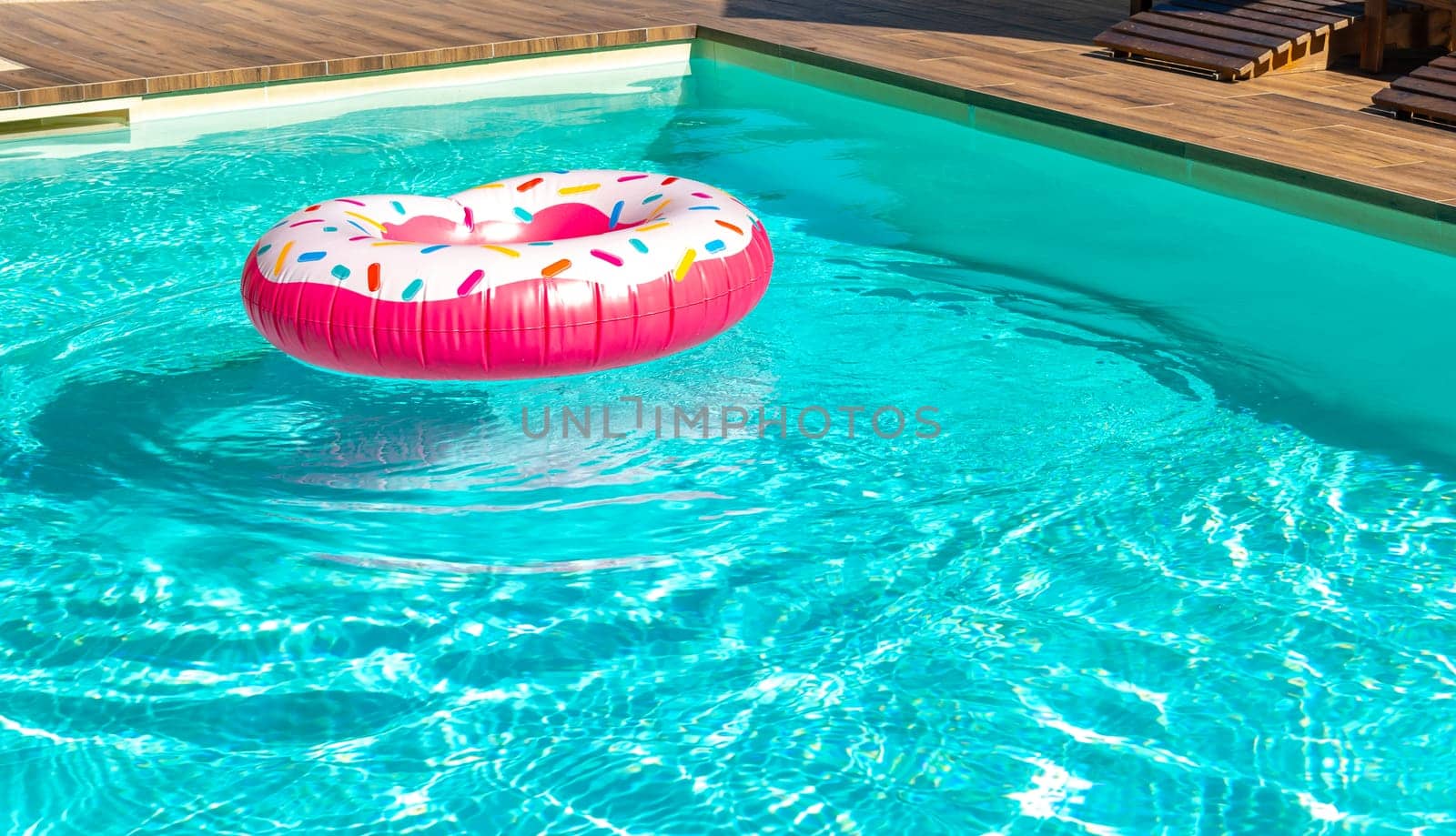 Pink inflatable donut-shaped float in turquoise swimming pool with wooden deck in sunny backyard