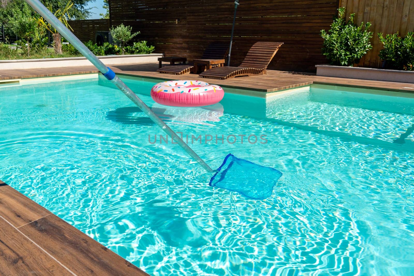 Skimmer net cleaning turquoise swimming pool water with wooden deck and colorful inflatable ring