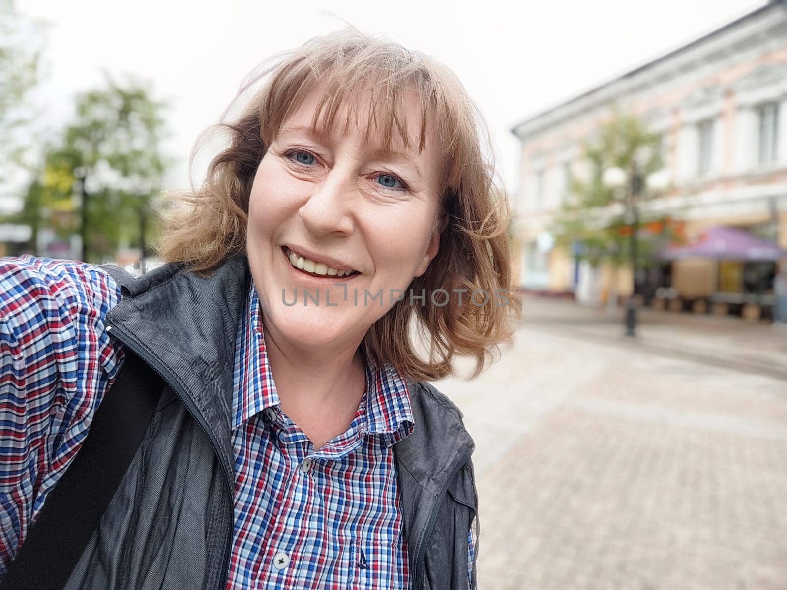 A middle-aged blonde woman enjoys a fun moment taking a selfie while exploring a quaint city street.