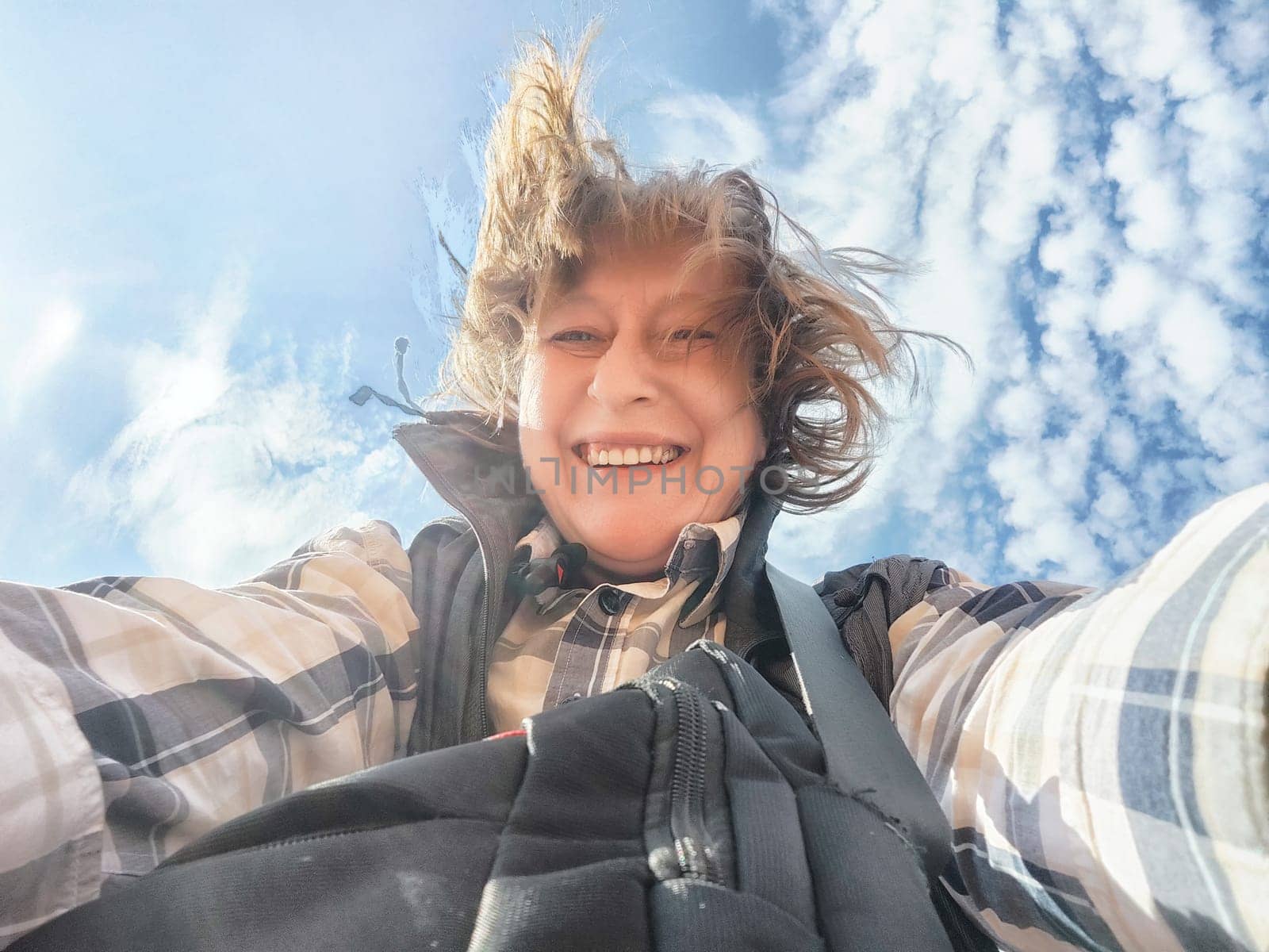 Middle-aged female travel blogger smiles for a selfie while exploring nature under a bright blue sky.