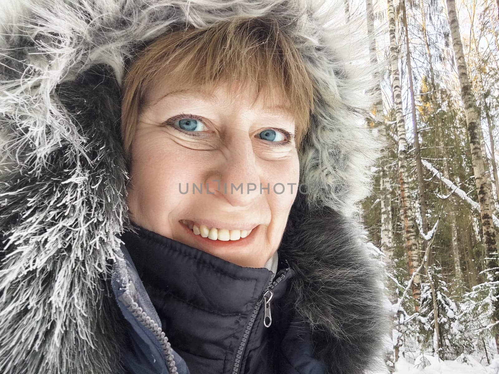 A mature blonde woman takes a cheerful selfie in a snowy forest landscape, dressed warmly for the cold weather.