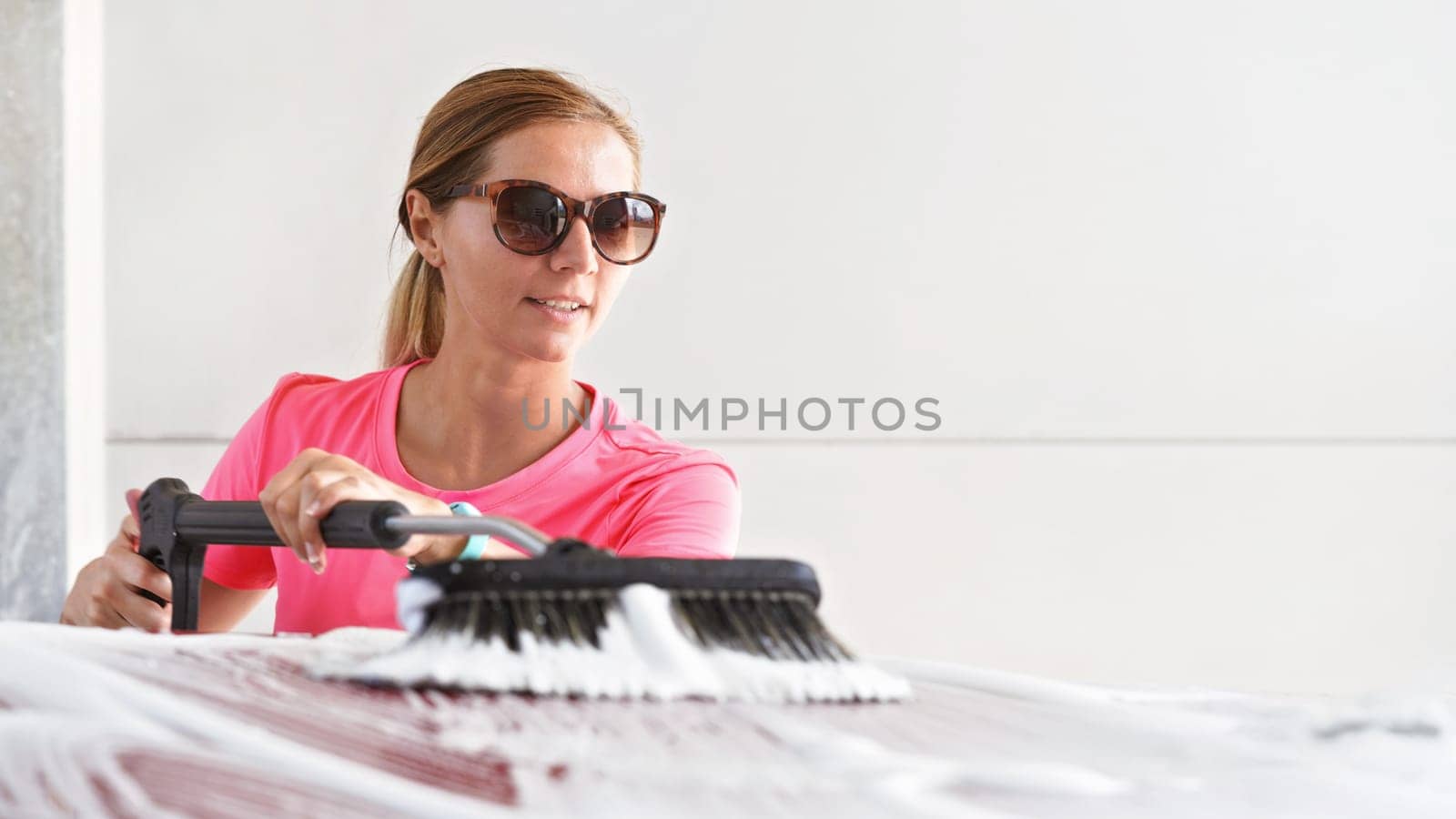 Young woman wearing pink t shirt and sunglasses cleaning her car in self serve carwash, spreading foam with brush over roof, banner with space for text right side