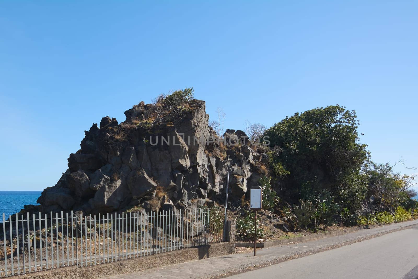 Scenic travel destination. A pedestrian sign located along a picturesque coastal road in Catania, Sicily, Italy. Surrounded by blooming flowers and a clear blue sea in the background.