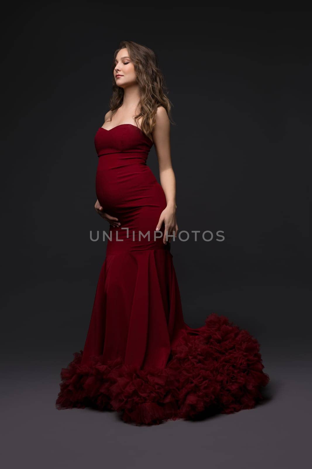 Serene pregnant female wearing long red dress standing with closed eyes on gray background and touching tummy