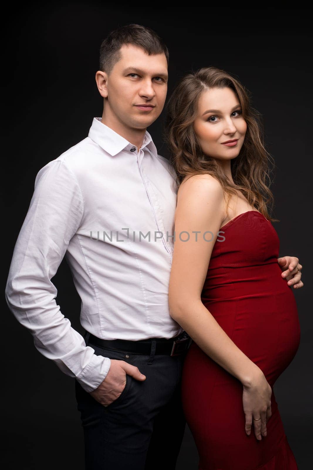 Handsome young man in classy outfit hugging beautiful pregnant wife in elegant red dress and looking at camera while standing against black background