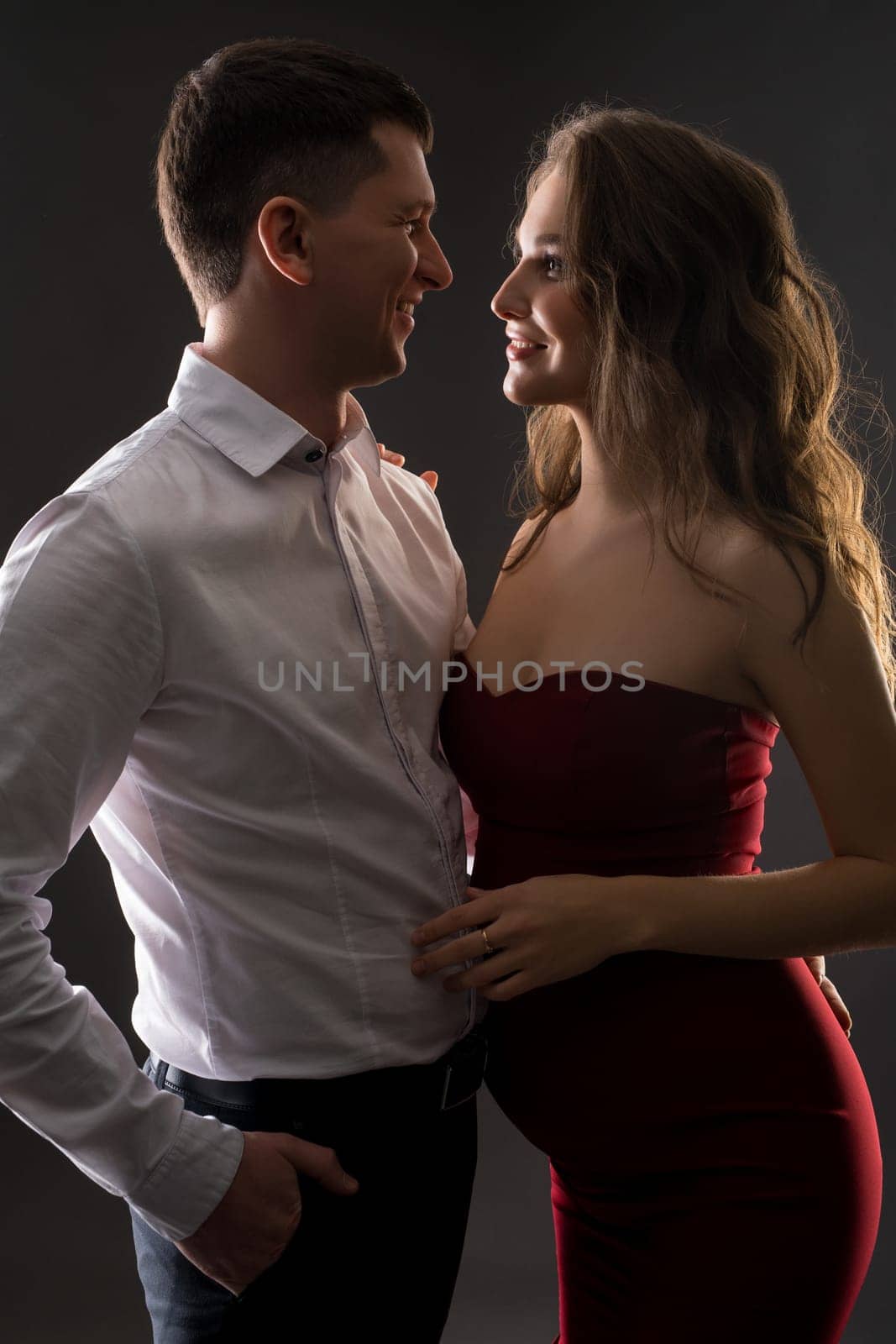 Handsome young man in classy outfit hugging beautiful pregnant wife in elegant red dress and looking one each other while standing against black background