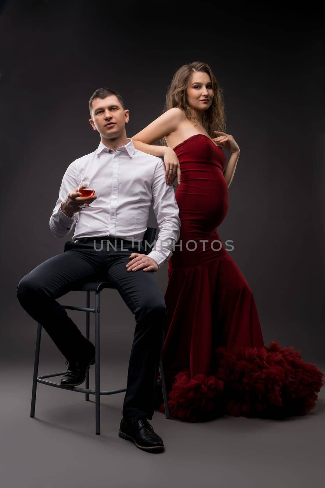 Pregnant woman in elegant red dress and man in stylish clothes looking at camera on gray background in studio