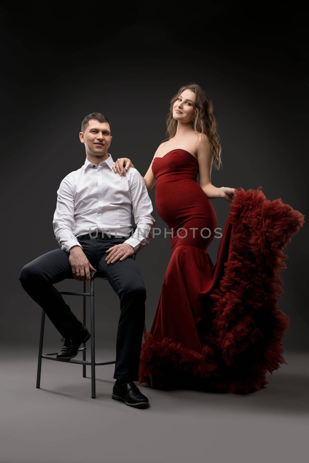 Full body of charming young pregnant female in elegant red gown standing near confident husband in studio with gray background