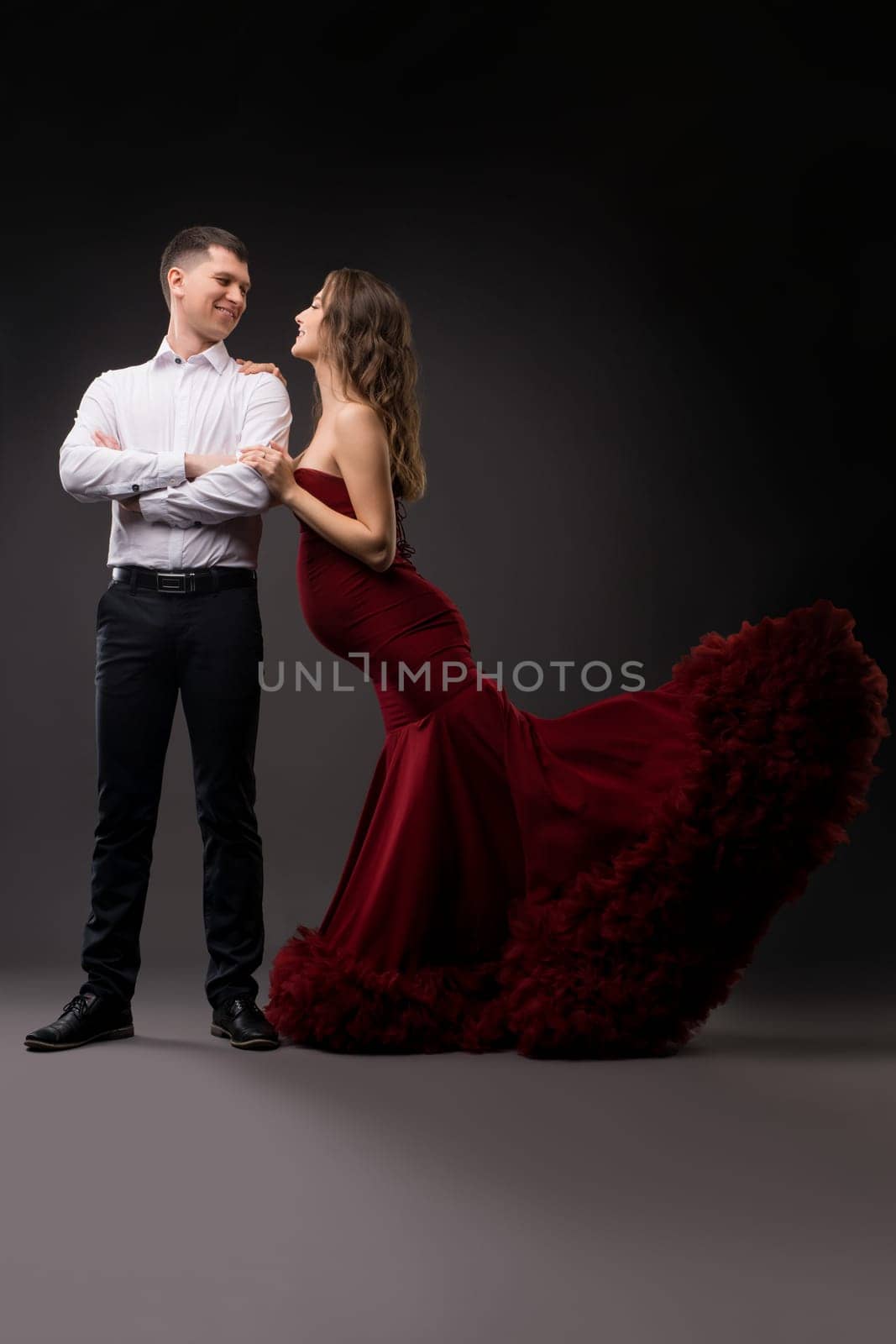 Full body of charming young pregnant female in elegant red gown standing near confident husband in studio with gray background