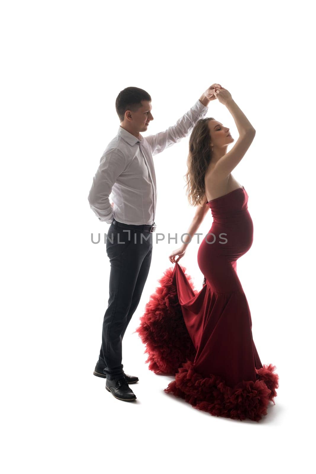 Side view of elegant man dancing with pregnant woman in red maxi dress on white background in studio