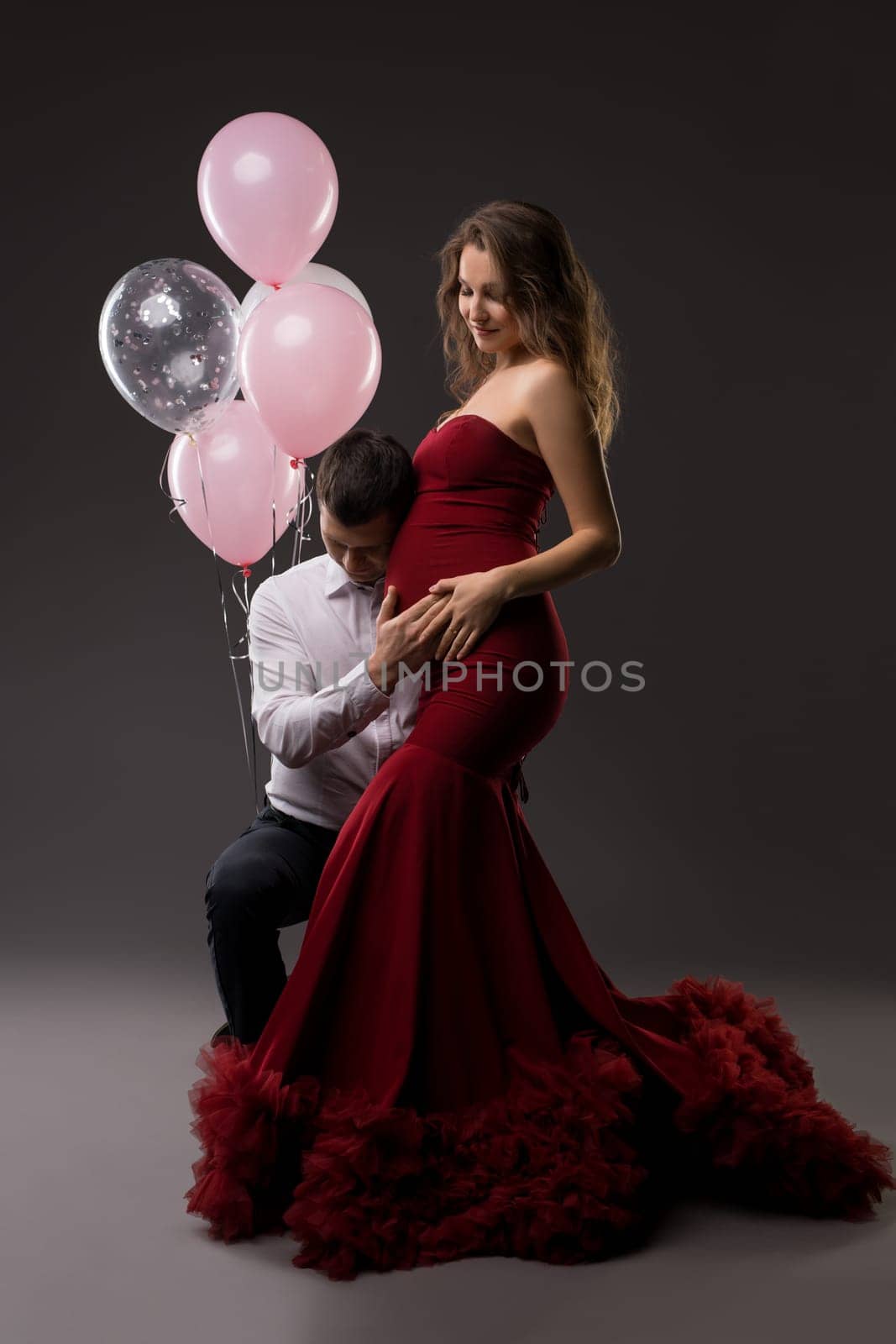 Side view of happy young man in elegant outfit hear belly from pregnant wife - stylish dress while standing together - dark studio decorated with pink balloons