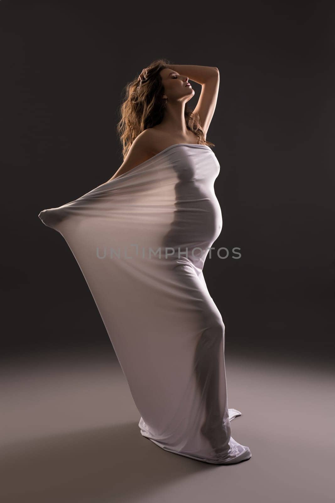 Sensual nude pregnant female wrapped in white cloth standing on dark background in studio