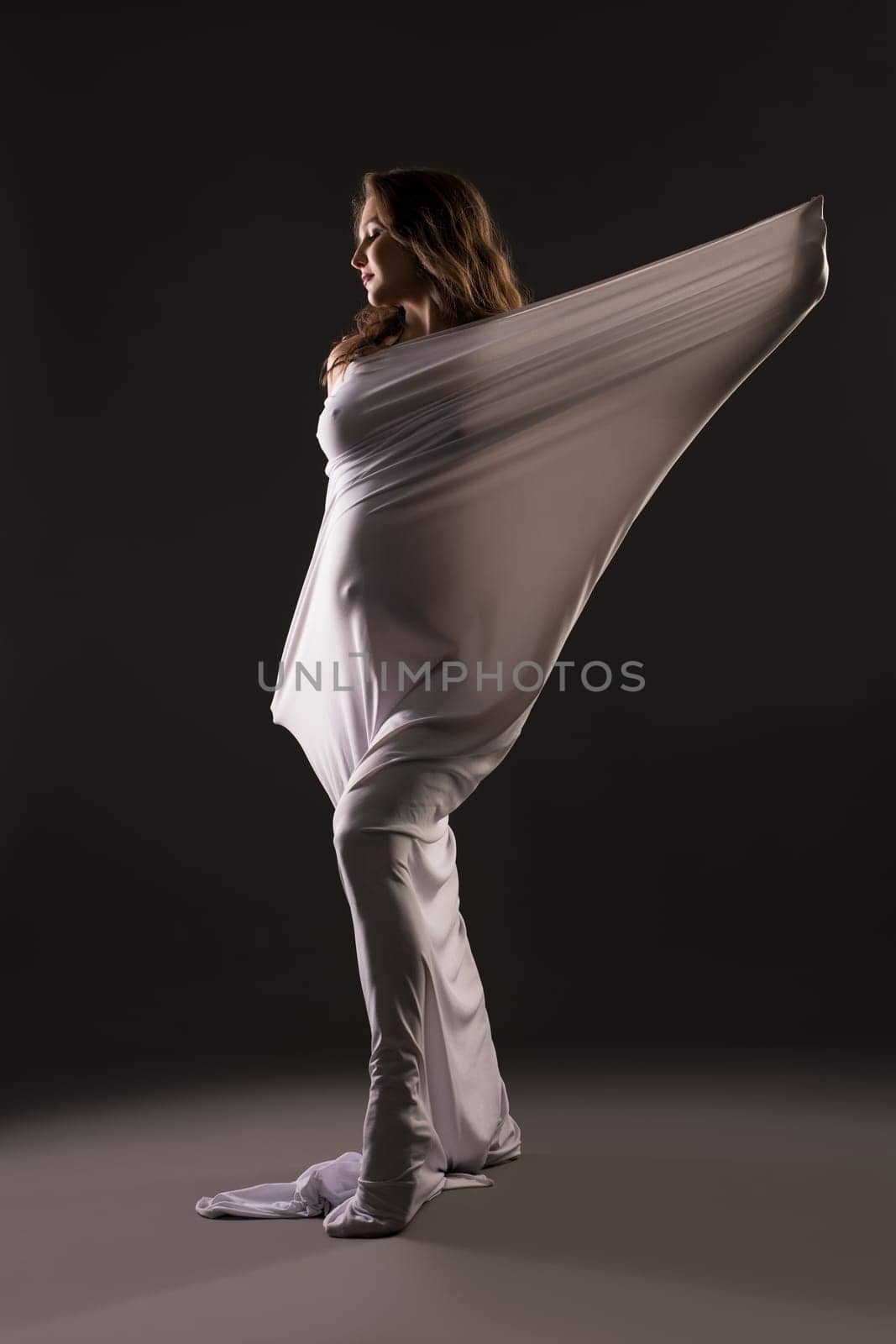 Sensual nude pregnant female wrapped in white cloth standing on dark background in studio