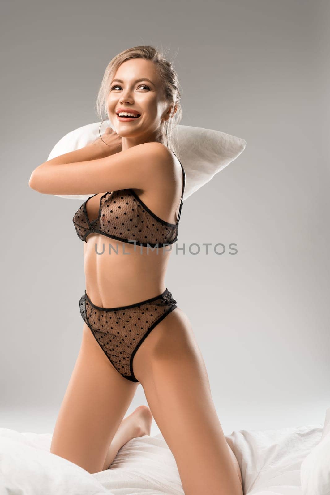 Alluring young blond woman in lace transparent bra and panties in polka dots sitting on bed and fighting pillow on gray background